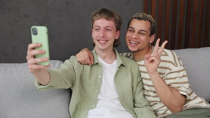 Young couple two gay men sit on sofa couch make selfie photo on mobile cell phone stay at home hotel flat rest relax spend free spare time in living room indoor Pride day june month love lgbtq concept - Powered by Adobe