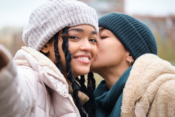 Happy dominican lesbian couple taking a selfie while kissing on the cheek at street. - obrazy, fototapety, plakaty