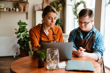 Young woman using laptop while training man with down syndrome to work in cafe
