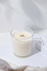 White aroma candle in glass jar with wooden wick on white background with books and cloth
