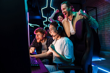 Group of friends celebrating victory of asian girl in video game while standing next to her in cybersport club