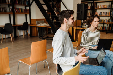 Smiling man and woman talking and using laptops while studying in library