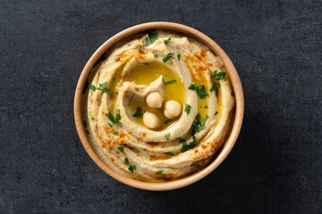 Chickpea hummus in a wooden bowl garnished with parsley, paprika and olive oil on black slate background. Top view