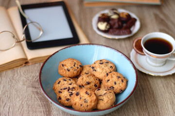 Books, reading glasses, e-reader, plate of chocolate pralines, bowl of cookies, cups of tea and lit candles on the table. Selective focus.