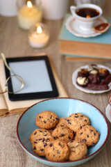 Books, reading glasses, e-reader, plate of chocolate pralines, bowl of cookies, cups of tea and lit candles on the table. Selective focus.