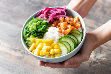 Hands showing a poke bowl with rice, salmon,cucumber,mango,onion,wakame salad, poppy seeds ands...