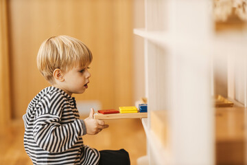 A boy is putting educational toy on the shelf after playing with it in kindergarten.