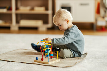 A little boy is sitting on the floor at kindergarten and learning logic and motor skills at kindergarten by playing with educational toy. - obrazy, fototapety, plakaty