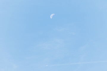 The waning moon is in the daytime in the blue sky underneath a commercial airliner is flying