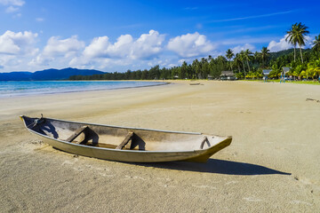 Small Indonesian traditional wooden ship or Jongkong lying on the Tropical beach with coconut trees, traditional house, and blue sky with clouds on Sunny day.