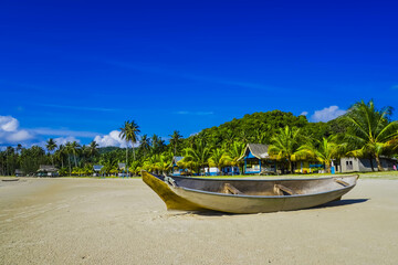 Fototapeta na wymiar Small Indonesian traditional wooden ship or Jongkong lying on the Tropical beach with coconut trees, traditional house, and blue sky with clouds on Sunny day.