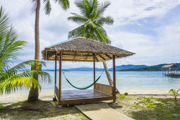 Tropical summer beach resort with palm, coconut trees, and hammock in Piugus Island, Anambas, Indonesia on a beautiful summer day with colorful ocean waters, pristine sands and cloudy sky. Copy space.