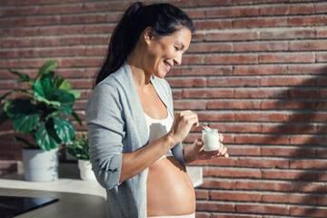 Beautiful cheerful pregnant woman with braces eating a yogurt in the kitchen at home