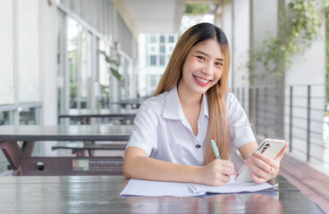 Obraz premium Young Asian woman student in uniform using smartphone and writing something about work. Documents on the table at university in Thailand. Her face with smiling in a working at to search information.