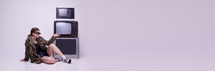 Young girl in stylish animal print coat and sunglasses sitting near retro TV sets against grey...