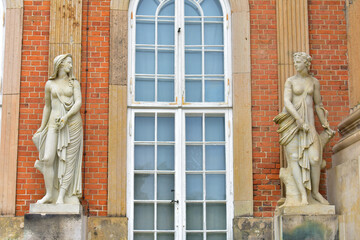 Antique statues of two women on a pedestal at the facade of the building. Brick red wall and tall old window in a white frame. Monumental art. Germany, Potsdam, Royal Castle, August 2022.