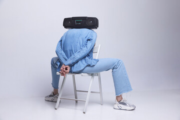 Young girl in jeans clothes sitting on chair with retro music player on her back against grey...