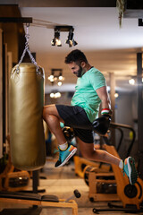 Muscular man with boxing gloves jump hitting a punching bag with a knee in a fitness studio.