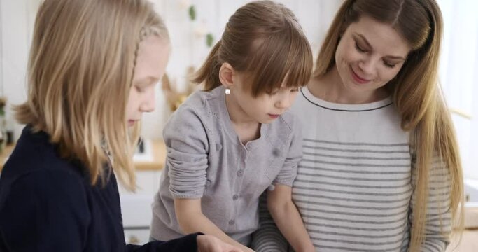 Happy young mother talking and assisting cute daughters in making fresh cookies with dough in kitchen during weekend Generative AI