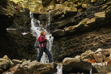 Obraz premium Hiking in mountains. Woman enjoying hike on sunny vacation day. Female with backpack walking close to waterfall. Spending summer vacation close to nature