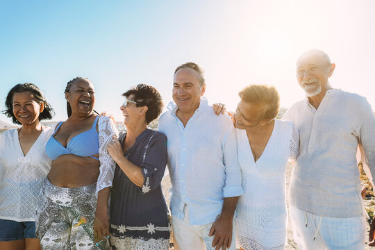Happy senior people having fun walking on the beach at sunset wearing summer clothes - Joyful elderly lifestyle, vacation and travel concept - Main focus on center friends faces