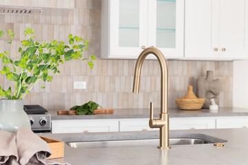 A kitchen faucet detail with a grey stone countertop island, gold faucet, stainless steel...