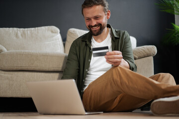 Happy, smiling customer sitting on floor next to sofa while shopping online using credit card for...
