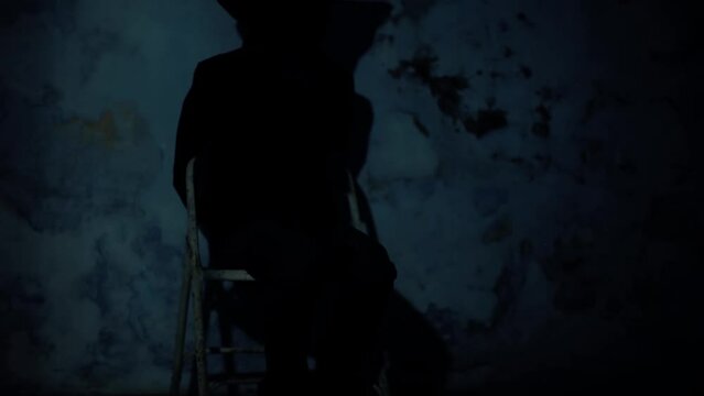 Cinematic shot of a man sitting on a chair with a black colored cloth covering his face, and his hands tied. Dark dramatic light. Hd footage.
