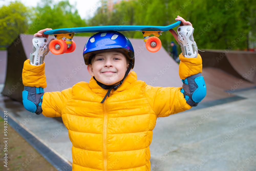 Wall mural portrait of a smiling schoolboy in a safety helmet with a bright blue skateboard, an urban cruiser in his hands in an outdoor skate park in spring, autumn