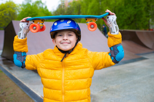 portrait of a smiling schoolboy in a safety helmet with a bright blue skateboard, an urban cruiser in his hands in an outdoor skate park in spring, autumn