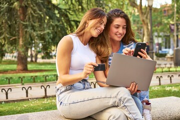 Friends shopping online with laptop and credit card in park.