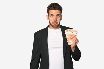 Young business caucasian man holding euro banknotes shrugs shoulders and open eyes confused.