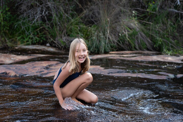 A happy girl in her swimming costume sitting in shallow river with water sliding over rocks during a family vacation