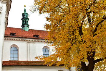 View of the old catholic church with white walls, red tiled roof and green tower, and a tree with yellow leaves. Autumn view, cloudy sky. Prague, Czech Republic, October 2022.