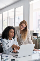 Vertical portrait of two businesswoman working together using laptop pc and talking about project. Small creative diversity team of African American and blond female executives meeting work in office. - obrazy, fototapety, plakaty