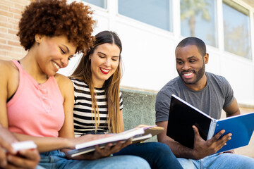 A group of young multi-ethnic students outside the university.The 3 young people sit happily looking at the book of the afro-haired girl to study for an exam.Concept of multiracial university students