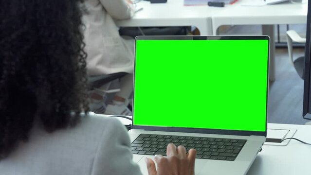 Young business woman working on laptop with blank copy space screen for ad app in modern busy office. Over shoulder view on businesswoman using touchscreen of computer with green chromakey mockup.