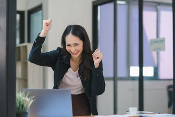 A businesswoman celebrates with her arms up while looking a laptop in a happy and successful pose.