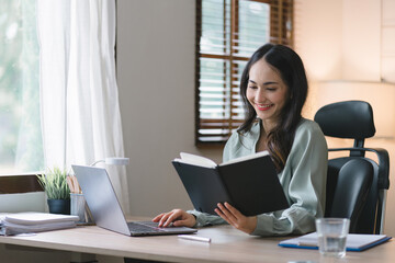 Fototapeta premium Image of a young, cheerful, happy, positive, cute, and beautiful Asian millennial businesswoman working on financial and marketing projects while sitting indoors in a home office.