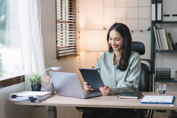 A cheerful mixed-race woman is using a laptop and a tablet for remote work or leisure while sitting indoors in a home office.