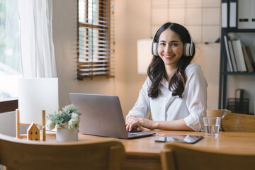 Image of a young, pleased, happy, cheerful, cute, and beautiful businesswoman sitting indoors at a dining table in her home office, using a laptop computer, and listening to music with earphones.