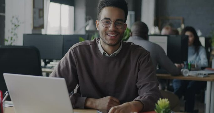 Slow motion portrait of joyful African American entrepreneur smiling sitting at computer desk in coworking space. Office work and employee concept. Generative AI