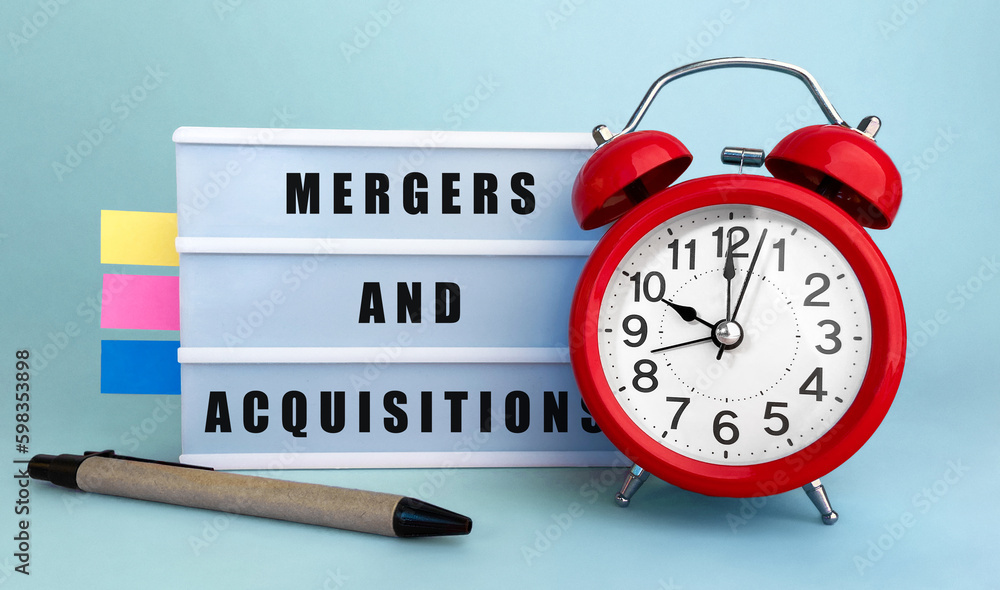 Poster text mergers and acquisitions written on the lightbox with alarm clock and colorfull stickers on blu