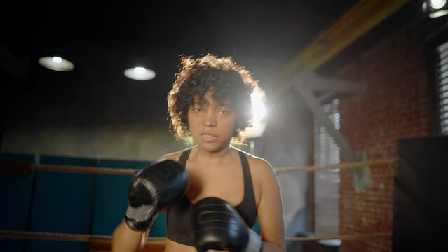 Afro american woman boxer in boxing gloves looks at camera stands on boxing ring and boxing. Sportswoman training in gym. Girl's power, woman train workout, sport fight, self-defense and feminism.