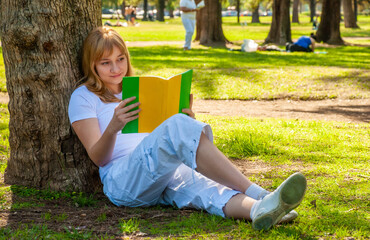 A girl with a book sits near a tree in the park