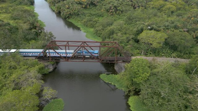 A historic train riding threw the jungle of Costa Rica over an old railway bridge, surrounded by rain forest, painted in Costa Rican flag colors (Aerial view, drone, 4K)