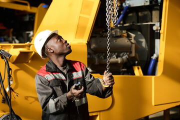 Young engineer in workwear and protective helmet looking upwards and pulling chain by hook while...