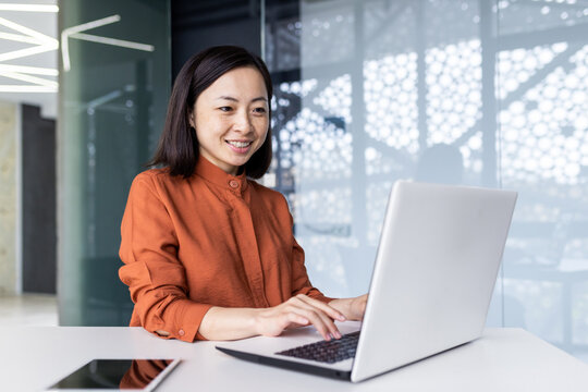 Successful asian business woman working inside office building with laptop, female worker smiling and looking at computer screen, satisfied with work achievement results.