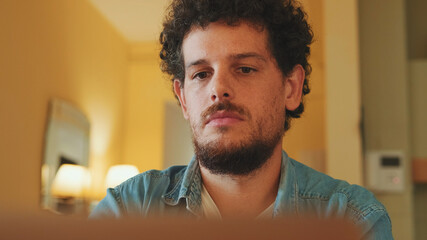 Close-up of guy dressed in denim shirt working on laptop while sitting in cozy kitchen