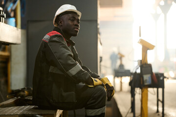 Young tired African American workman sitting on metallic bench while resting at break in the middle of working day in factory workshop